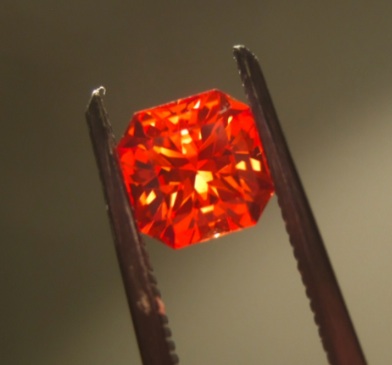 A small orange-red faceted stone held in tweezers.