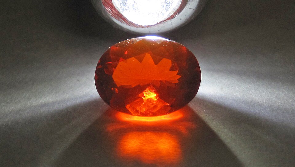 An oval orange-red fire opal on a gray surface, lit from above.