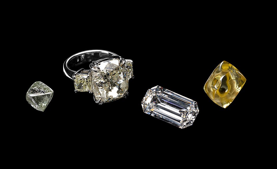 Four objects: a small diamond crystal, an old cushion in a ring, an emerald-cut rectangle, and a yellow crystal.